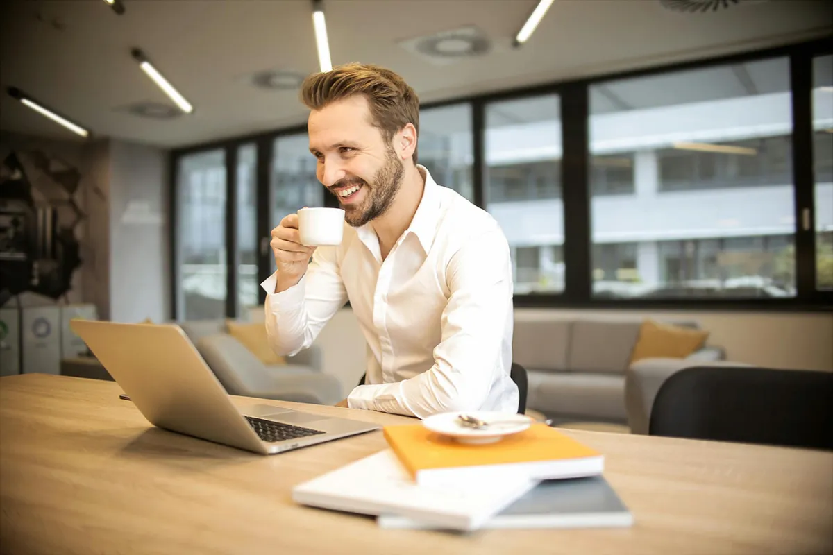 Smiling well-dressed and groomed man holding a cup of coffee while sitting at a table in front of a laptop.