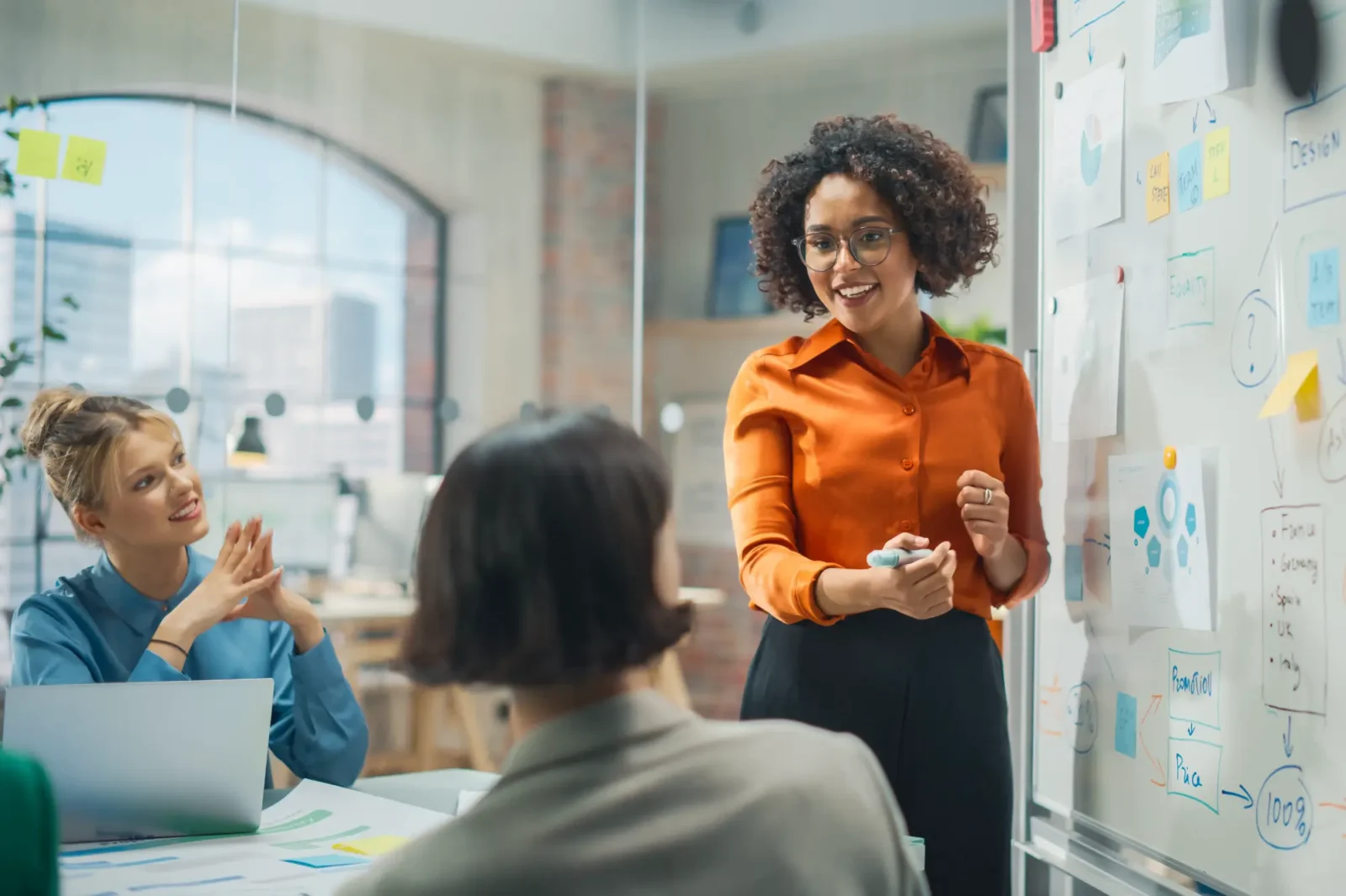 A young professional woman standing at a white board with charts while talking to colleagues sitting at a table.