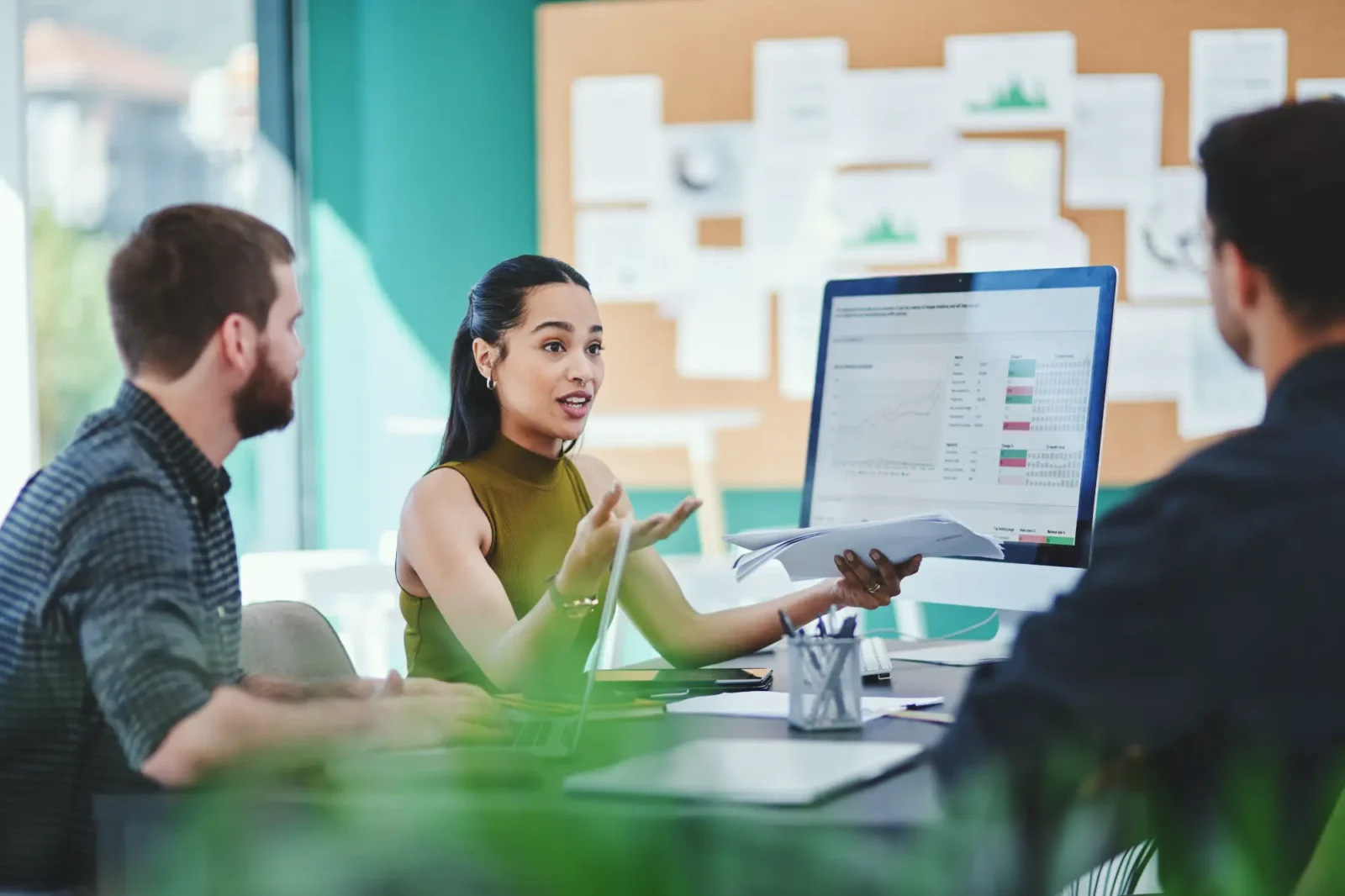 Business meeting with young woman speaking to colleagues with data charts on the computer screen and board behind them.