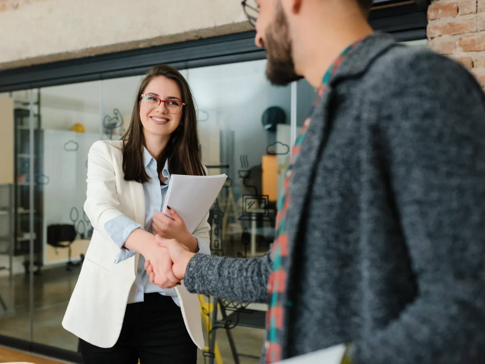 A professional young woman holding papers and shaking someones hand.