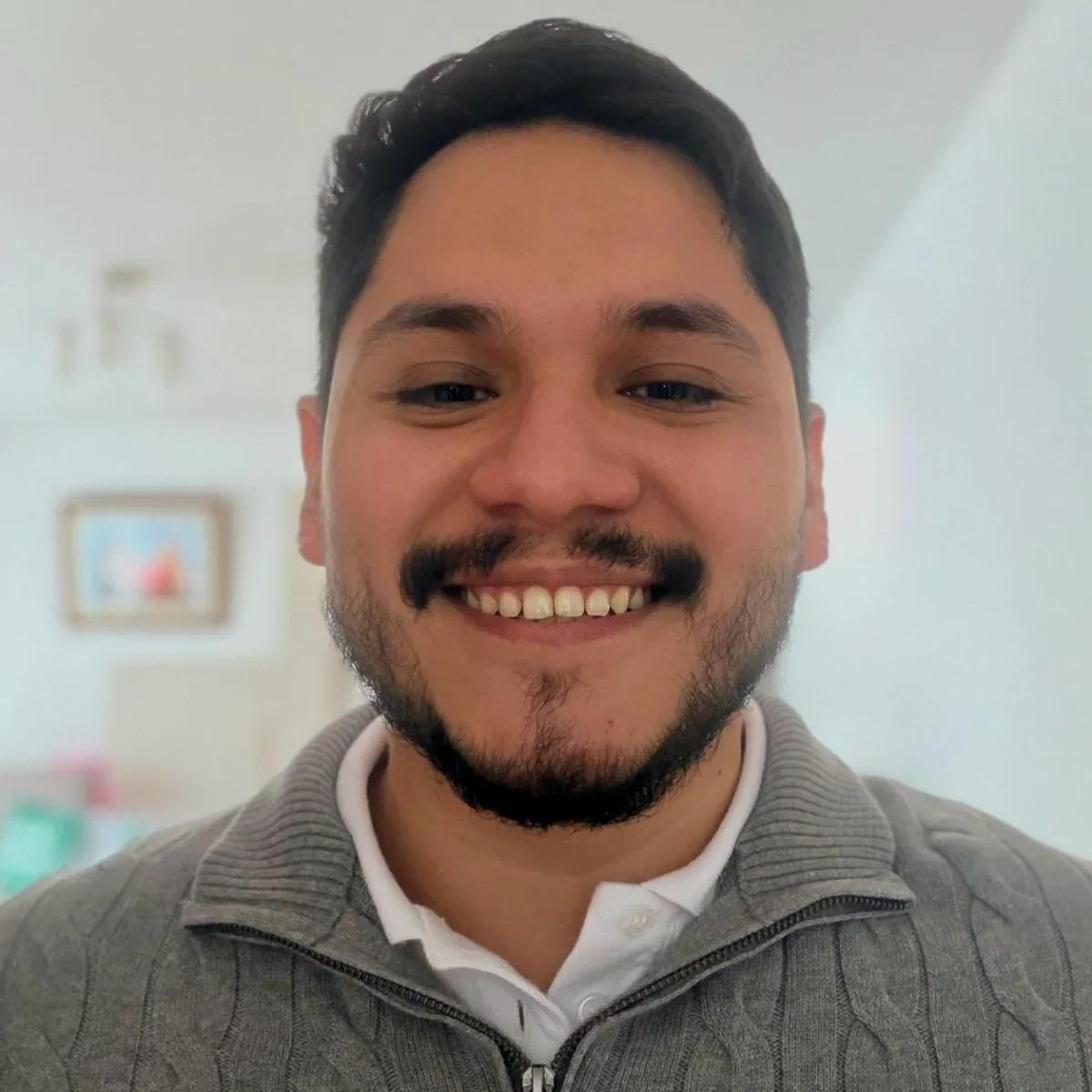 Cesar Huaman, with a beard and dark hair, smiles brightly while wearing a white shirt and a gray zippered knit sweater.