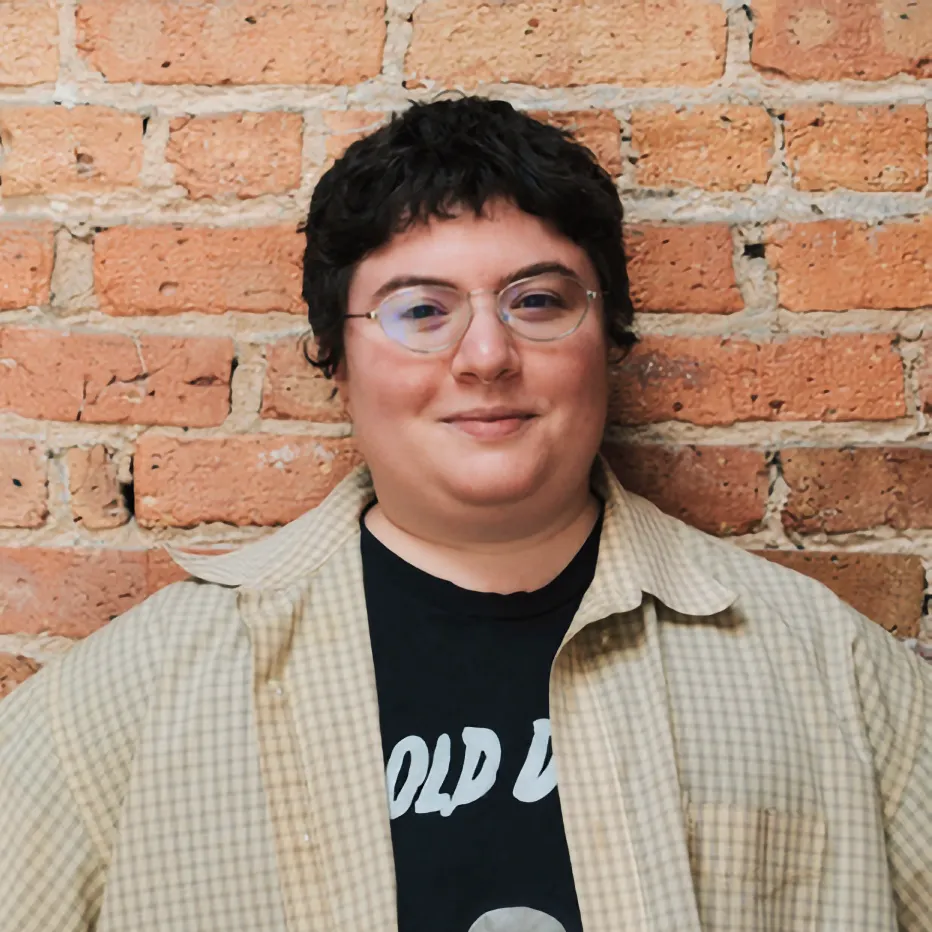 Elam Karabush, wearing glasses and a light checkered shirt, stands smiling slightly against an exposed red brick wall.