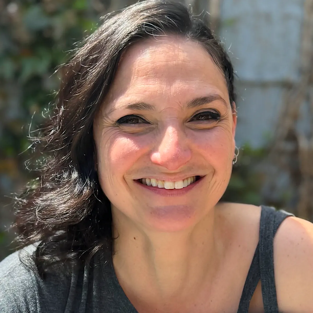 A close-up headshot of Jen Higgins smiling warmly. She has dark hair and is wearing a gray top, photographed outdoors in bright light.